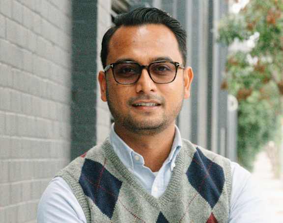 A man wearing glasses and a checkered vest stands with his arms crossed.