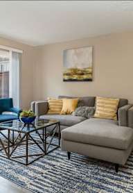 A living room with a grey couch, a blue chair, a coffee table, and a painting on the wall at Nola 624 Apartments, West Covina