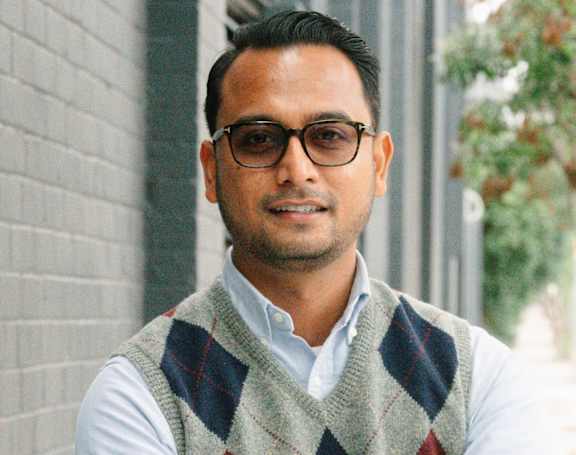 A man wearing glasses and a checkered vest stands with his arms crossed.