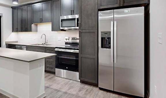 a modern kitchen with dark wood cabinets, light quartz countertops, a narrow central island, a tile backsplash, and stainless steel appliances