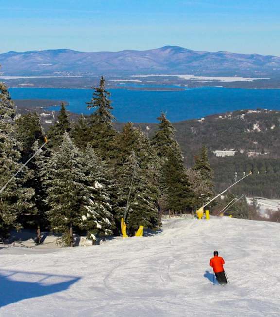 A skier is seen on a snowy slope with a lake and mountains in the background.