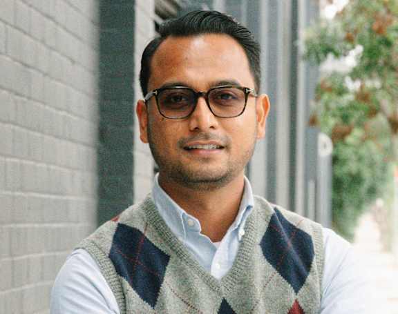 A woman with long dark hair wearing a red cardigan and black top standing in front of a window.