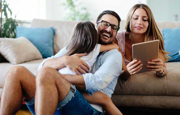A family of three is sitting on a couch with a tablet in hand.