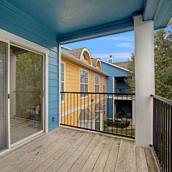 A wooden balcony with sliding glass doors and a gorgeous view of at the pathways leading to the blue and yellows apartments, surrounded by trees and grass at Woodland Park Apartment Homes. 