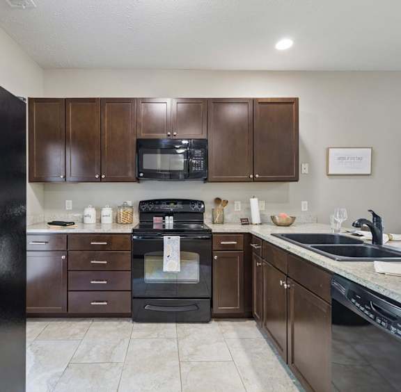  Staged apartment kitchen with dark wood cabinets, black appliances, speckled counters, and tile flooring.