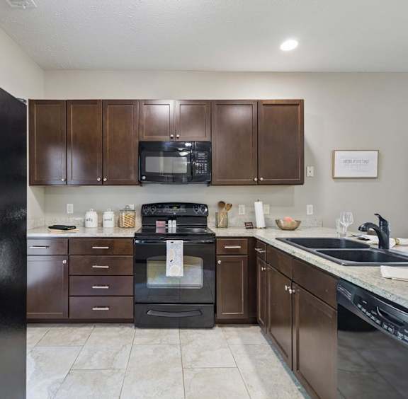  Staged apartment kitchen with dark wood cabinets, black appliances, speckled counters, and tile flooring.