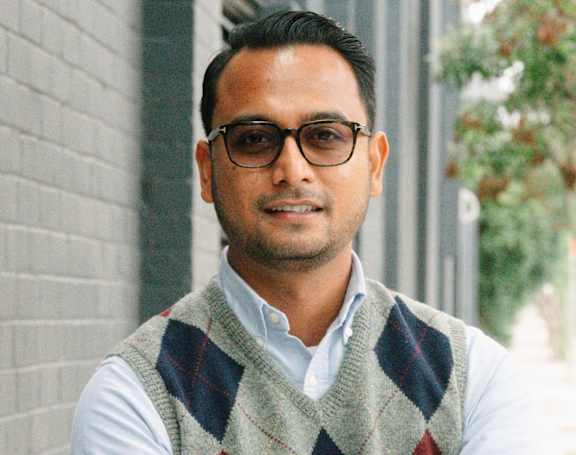 A man wearing glasses and a checkered vest stands with his arms crossed.