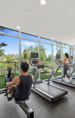 A man is working out on a rowing machine in a spacious gym with a view of the outdoors.