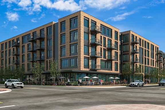 A large, modern building with many windows is surrounded by trees and a street with cars and pedestrians.