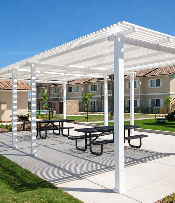 A white pergola with picnic tables is in the foreground of a playground.