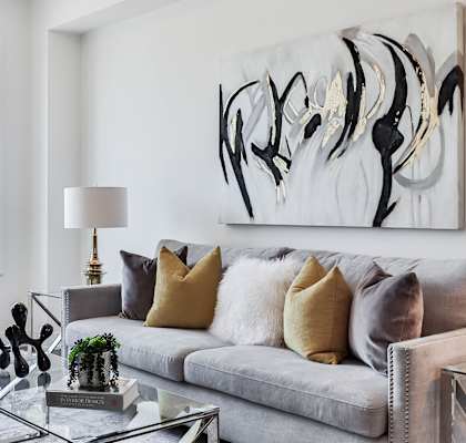 Living area with grey couch and colorful pillows.  Glass coffee table in front with black and white photo behind couch on wall.  White chair to left of couch and 2 windows in background.