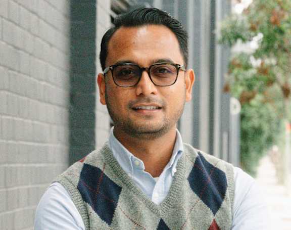 A man wearing glasses and a checkered vest stands with his arms crossed.