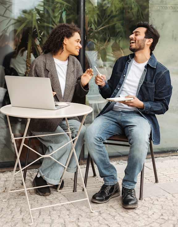 Two men are sitting at a table with a laptop and having a conversation.