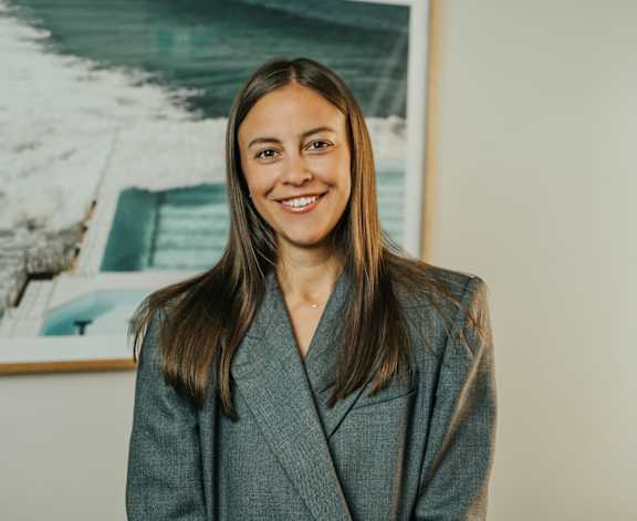A woman in a grey suit stands in front of a framed picture of a boat.