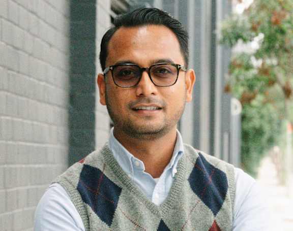 A man wearing glasses and a checkered vest stands with his arms crossed.