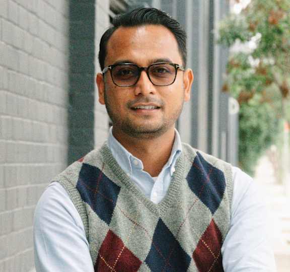 A man wearing glasses and a checkered vest stands with his arms crossed.