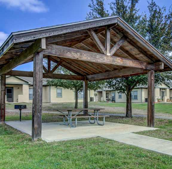 A wooden pavilion with a picnic table sits in the middle of a grassy area with houses in the background.