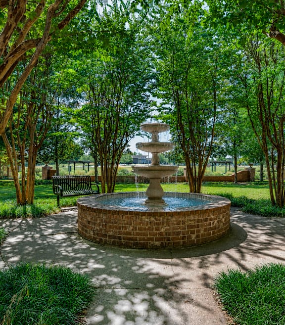 A circular brick fountain is the centerpiece of a park with benches and trees.