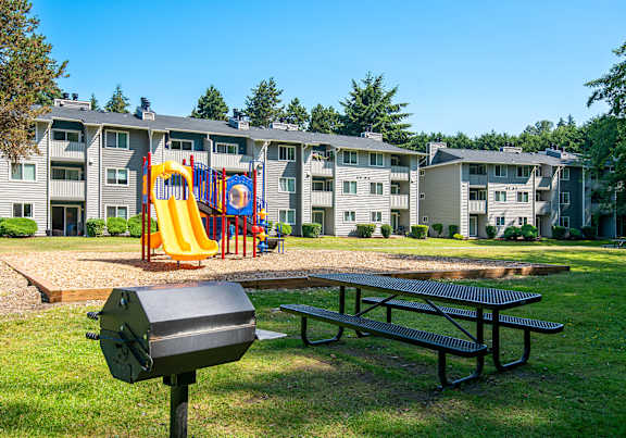 An exterior view of the community picnic table, barbeque grill, and playground with a slide at Pembrooke Apartments in Kent