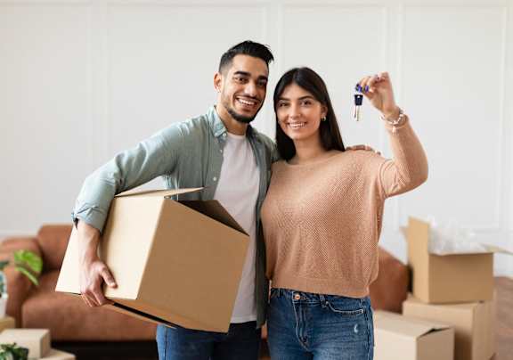 A man and woman are standing in a room with boxes, holding a key and a box.