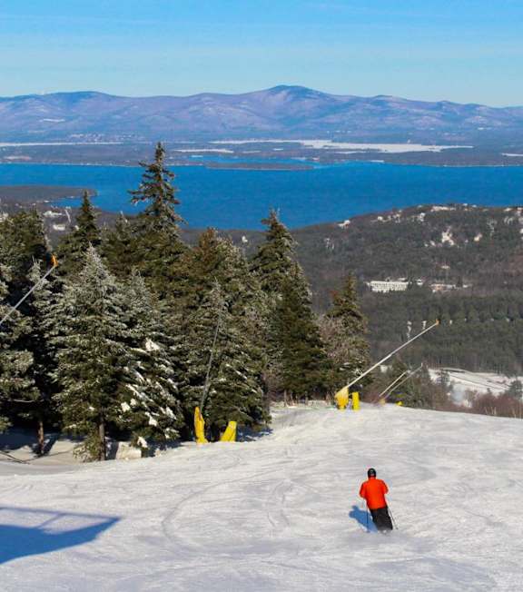 A skier is seen on a snowy slope with a lake and mountains in the background.