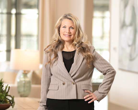 A woman in a beige blazer and black pants stands in a room with a wooden table and a potted plant.