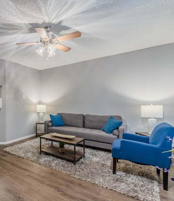 A living room with a grey couch, a blue chair, a dining table with chairs, and a ceiling fan.