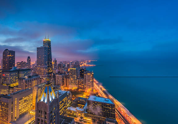 A cityscape at dusk with buildings illuminated and a body of water in the foreground.