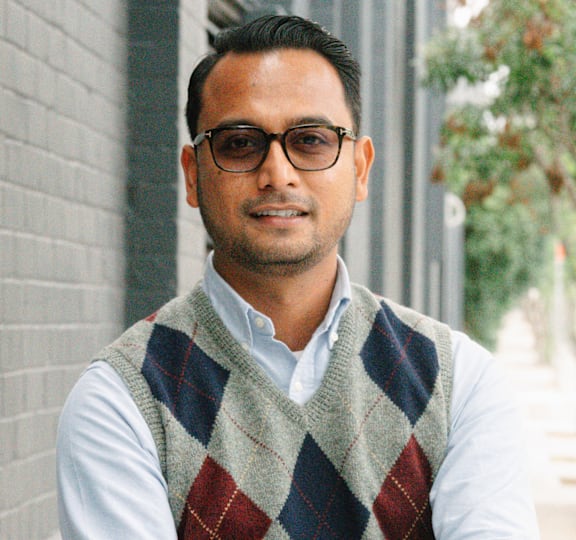 A man wearing glasses and a sweater vest stands with his arms crossed.