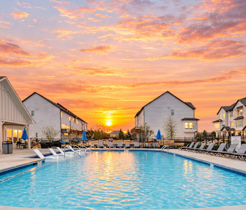 A swimming pool surrounded by lounge chairs and buildings under a sunset sky.