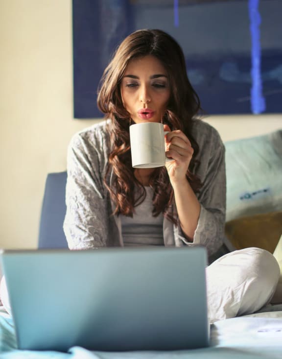 a woman sitting on her bed drinking coffee and looking at her laptop