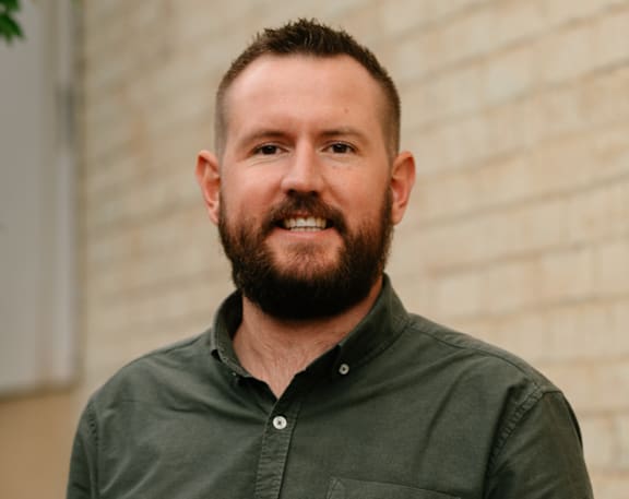 A man with a beard is standing in front of a brick wall.