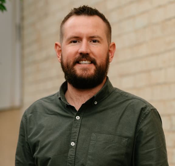A man with a beard is standing in front of a brick wall.