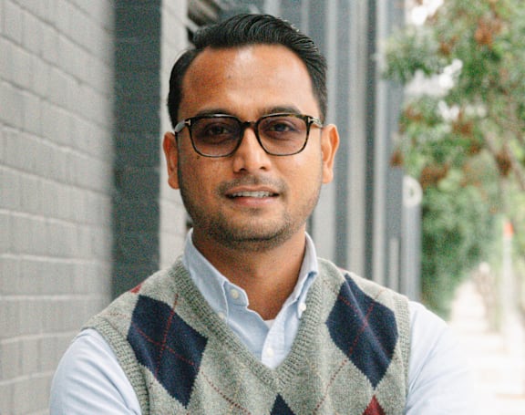 A man wearing glasses and a sweater vest stands with his arms crossed.