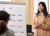 Thumbnail 10 of 10 - A woman stands in front of a whiteboard with writing on it in a classroom setting.