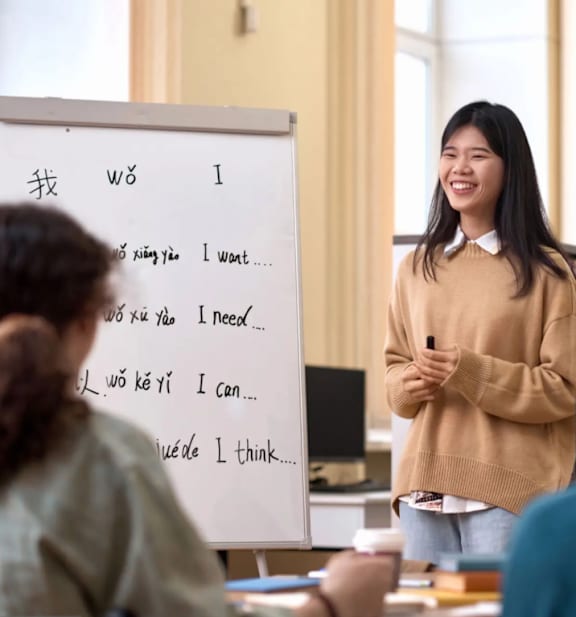 A woman stands in front of a whiteboard with writing on it in a classroom setting.