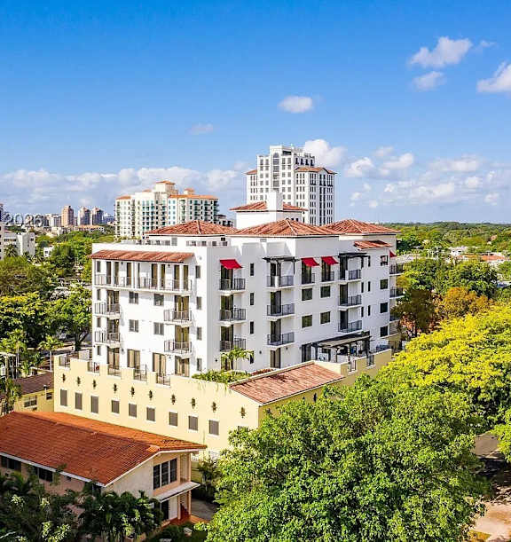 A white hotel with a red roof is surrounded by trees and other buildings.