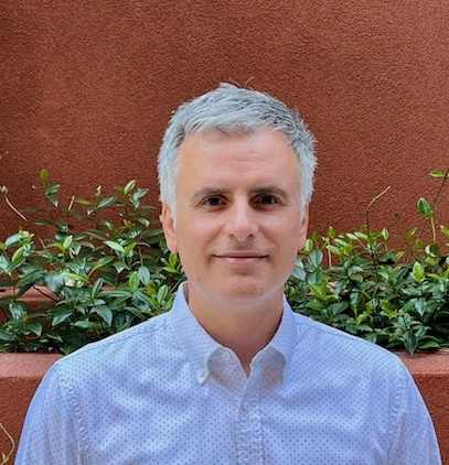 A man with grey hair wearing a light blue shirt stands in front of a red wall with green plants behind him.
