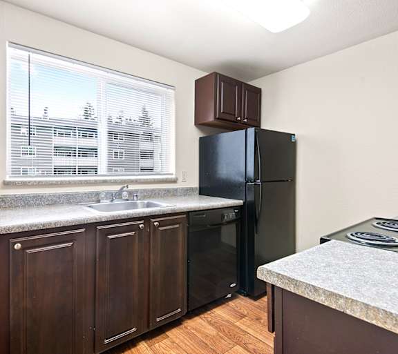 Kitchen view of the granite countertops, brown cabinetry, gas range, dishwasher, and fridge next to a sunlit window at Pembrooke Apartments in Kent