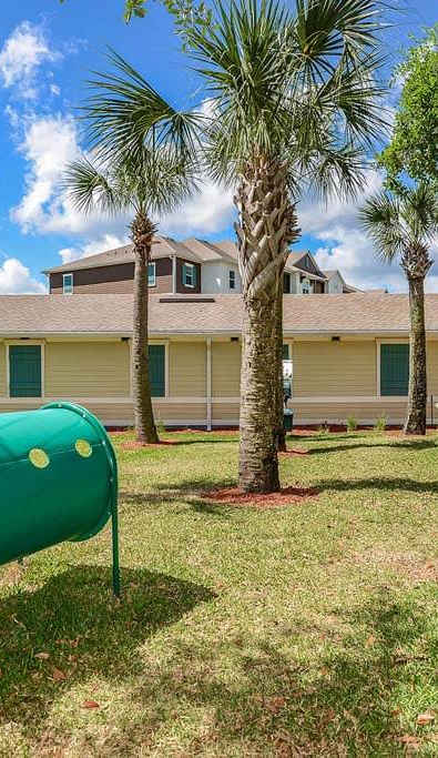 A green barrel is in the foreground of a grassy area with a playground and palm trees in the background.