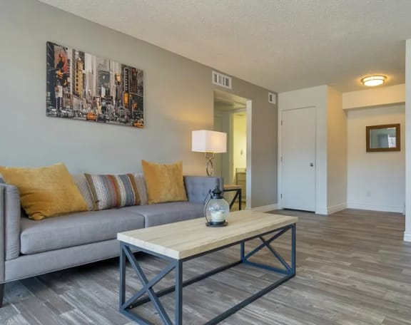 Cozy living room with plank flooring, a grey couch, and a wooden coffee table with metal legs at Spring Meadow Apartments in Glendale, AZ