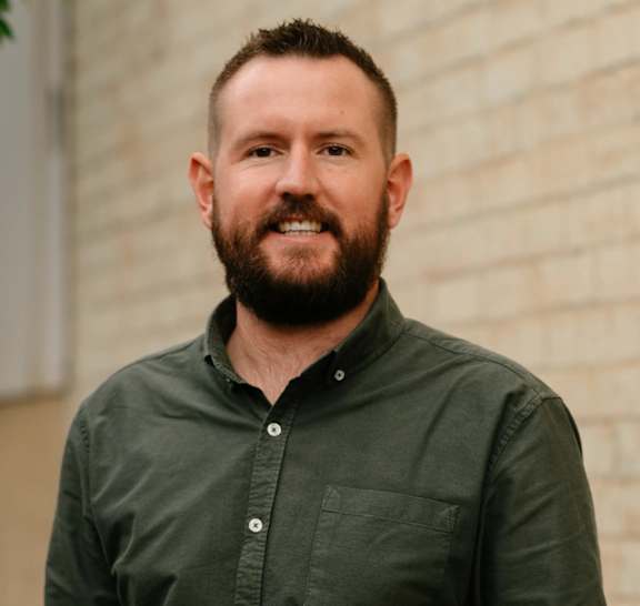 A man with a beard is standing in front of a brick wall.