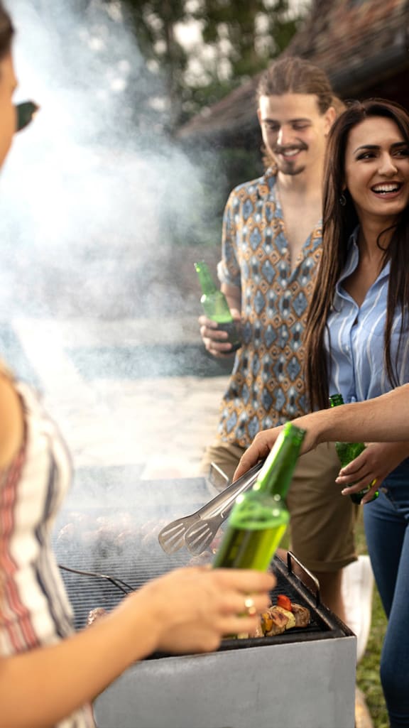 a group of people are gathered around a grill toasting bottles of beer