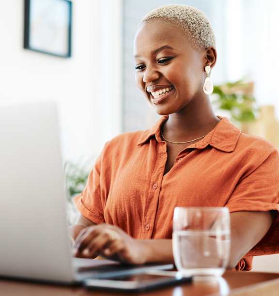 A woman in an orange shirt is smiling while using a laptop.