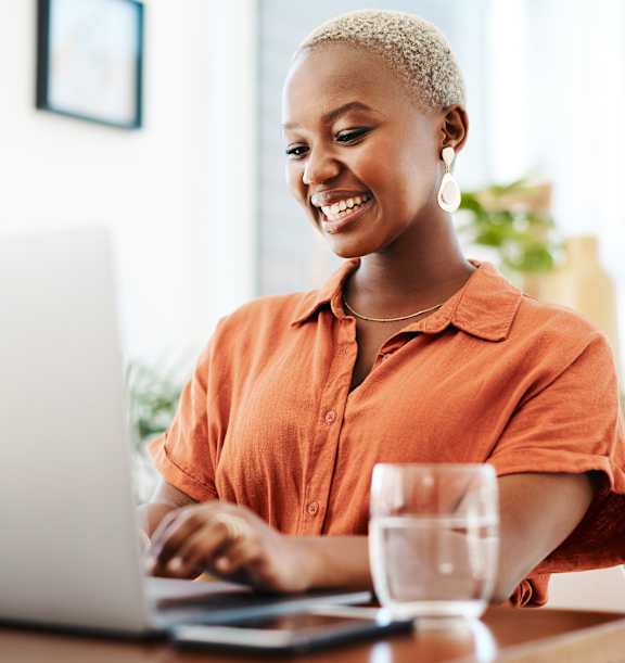 A woman in an orange shirt is smiling while using a laptop.