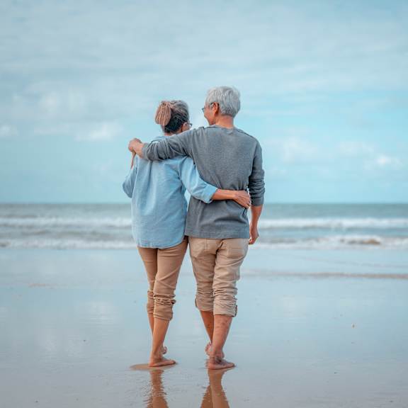 An elderly couple is walking on the beach holding hands.