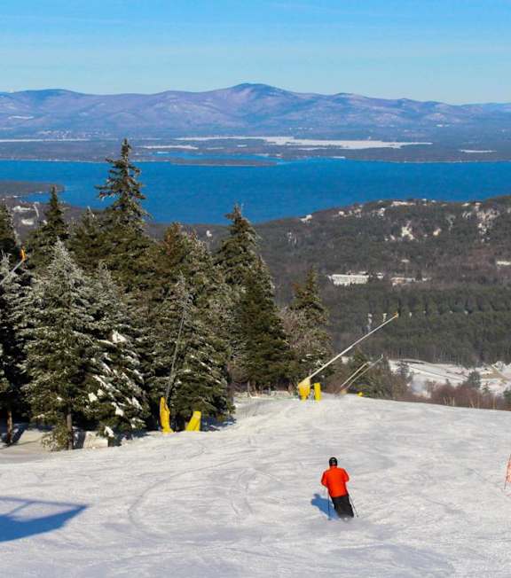 A skier is seen on a snowy slope with a lake and mountains in the background.