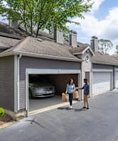 A man and woman are standing outside a house with a car in the garage.