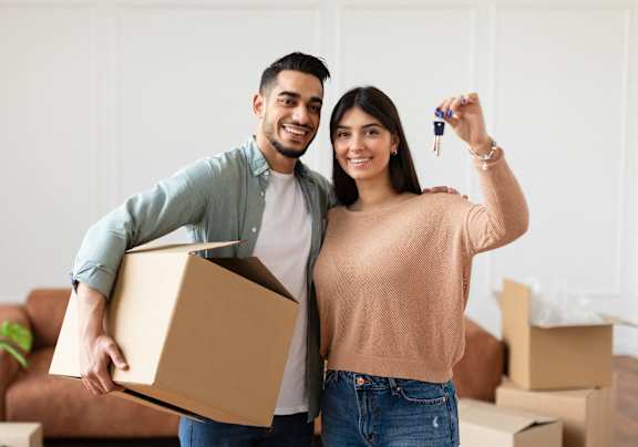 A man and woman are standing in a room with boxes, holding a key and a box.