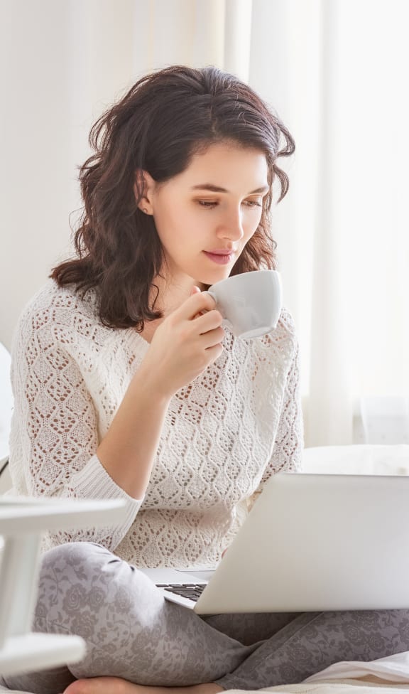 a woman sitting on her bed with a laptop and a cup of coffee